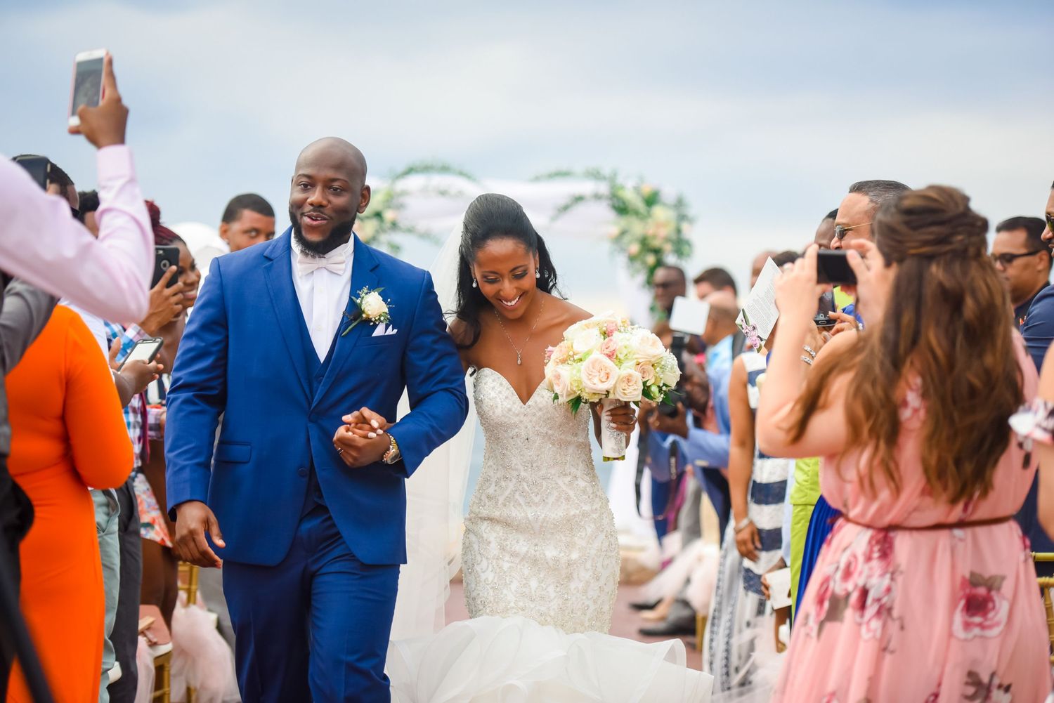 Wedding couple walks down the aisle as guests take photos during an outdoor ceremony.