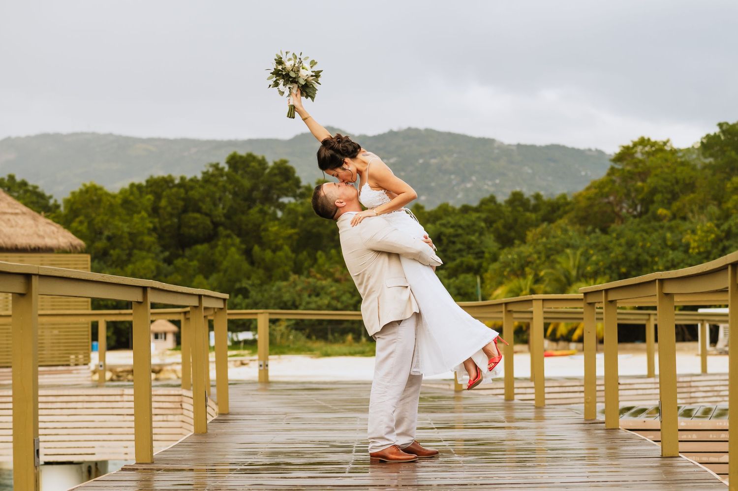 Playful wedding couple celebrates on wooden boardwalk with tropical scenery.