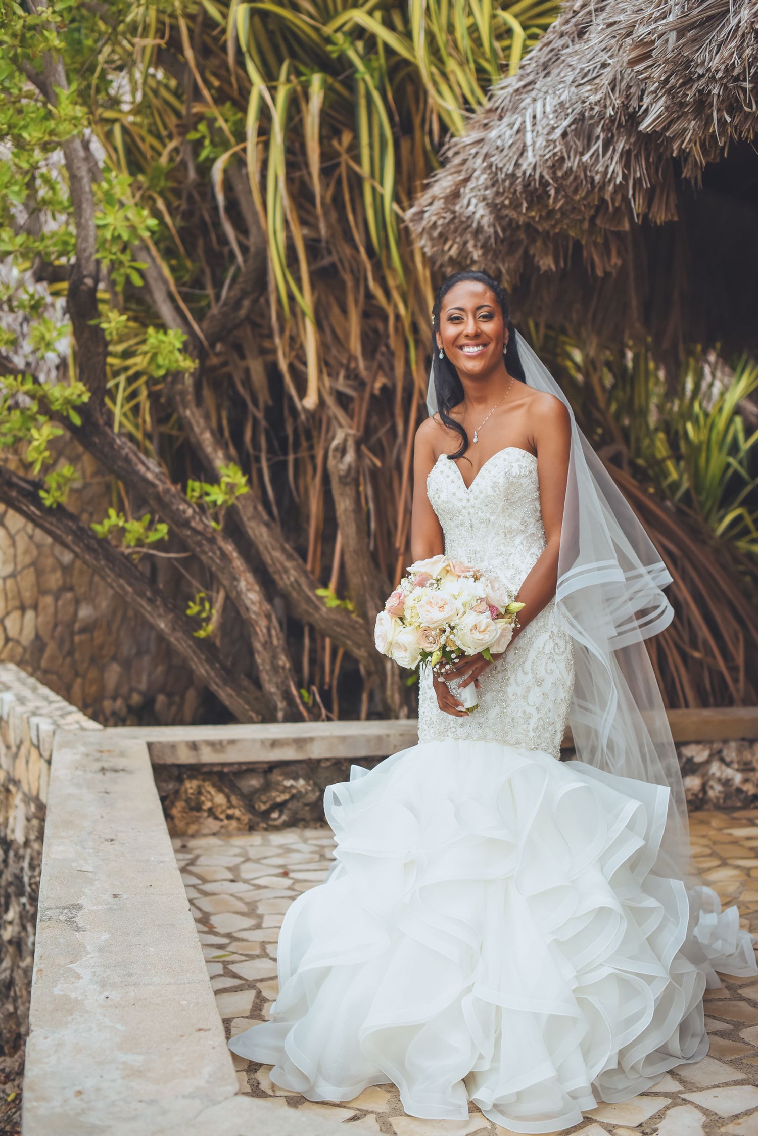 Bride wearing a ruffled ballgown sits gracefully on stone steps surrounded by tropical foliage.