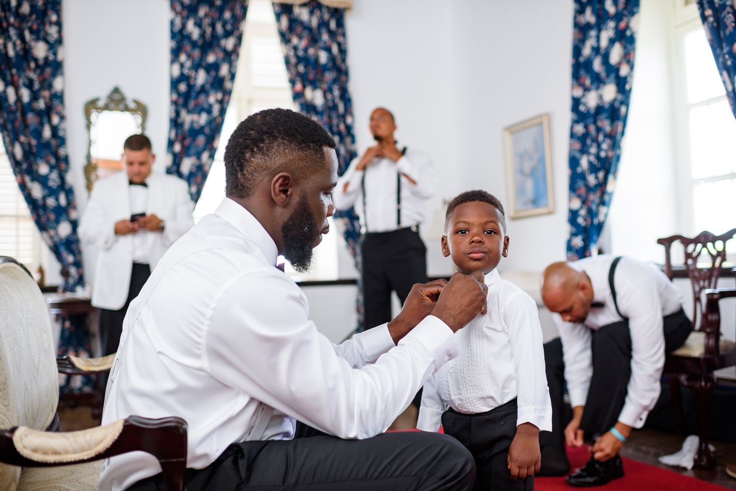 Wedding party members in white shirts preparing in a room with blue floral curtains.