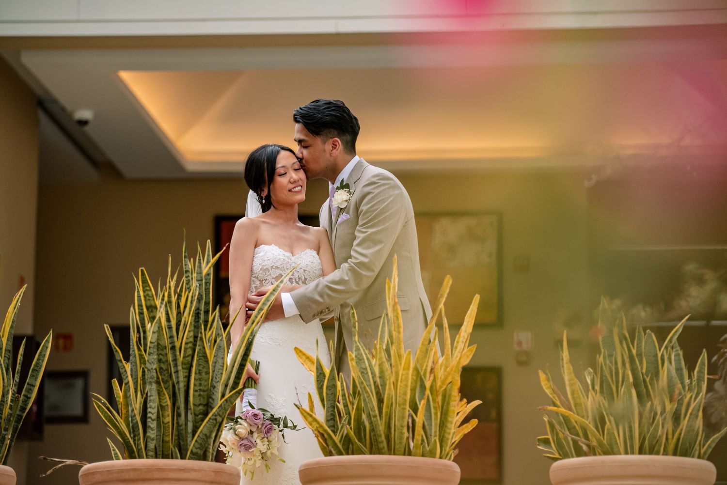 A romantic wedding photoshoot with a couple posing near decorative plants in an indoor venue.