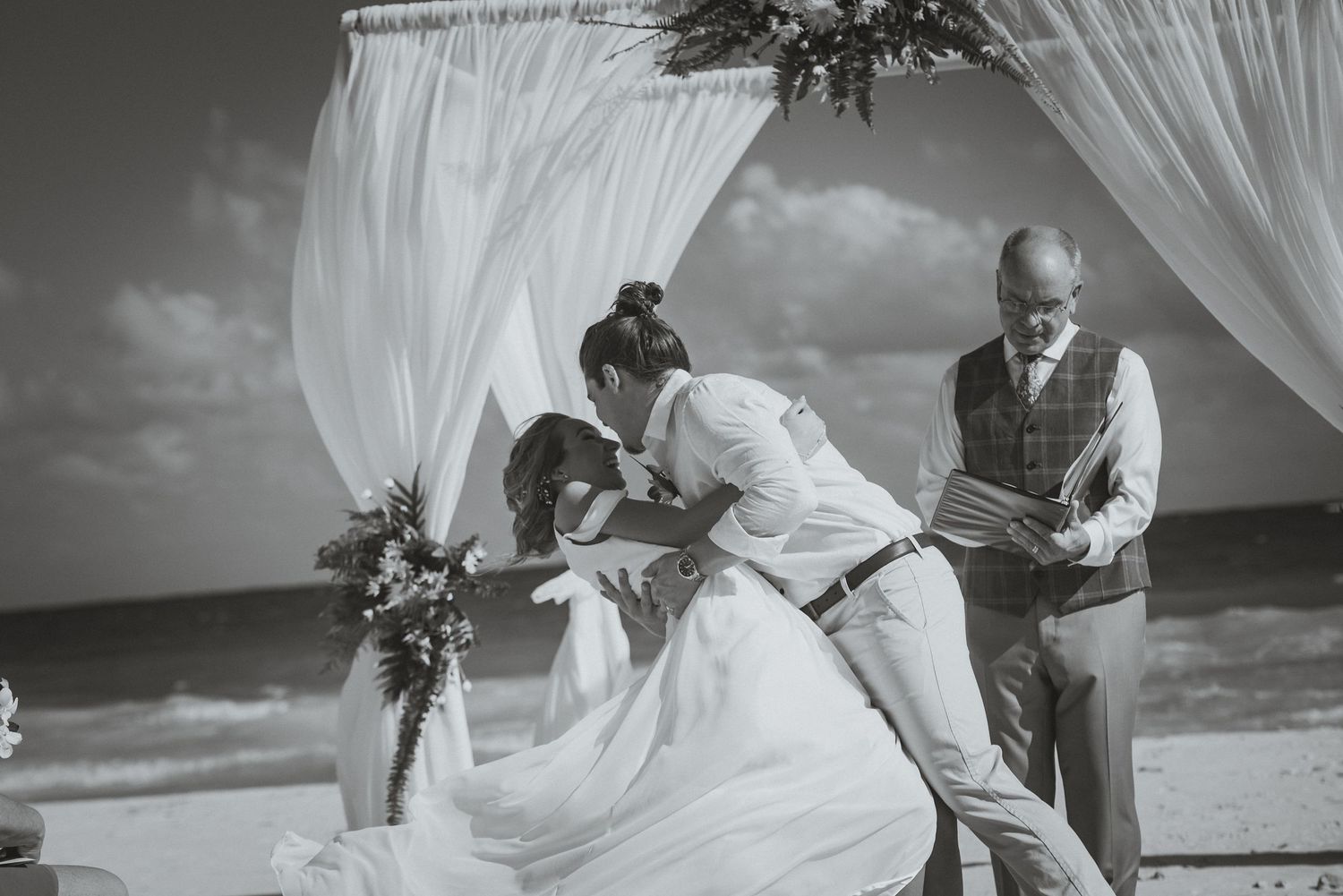 Wedding party poses joyfully on a scenic beach with white flowing curtains and tropical foliage in the background.