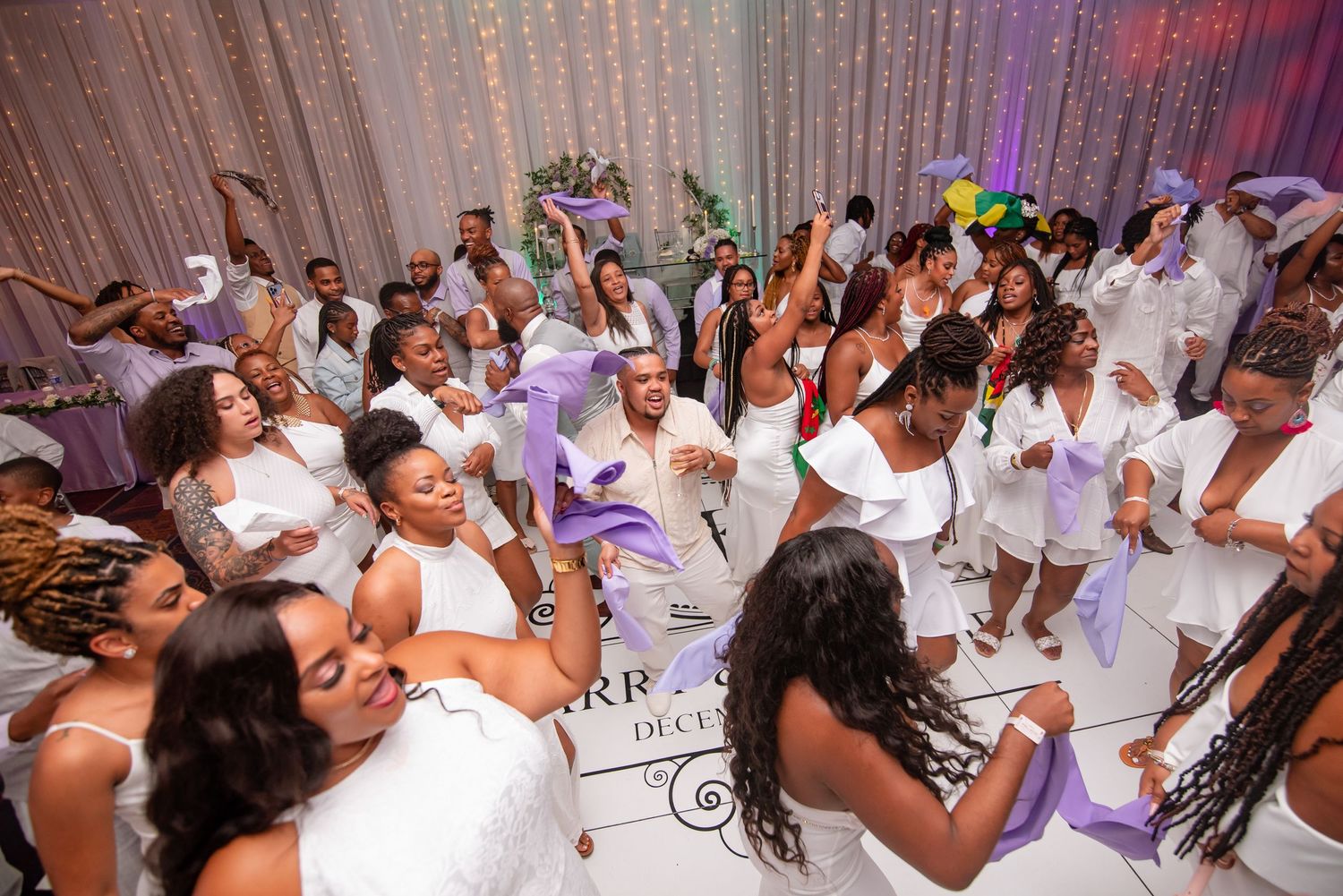 Wedding guests dancing and celebrating on a white dance floor with purple lighting accents.