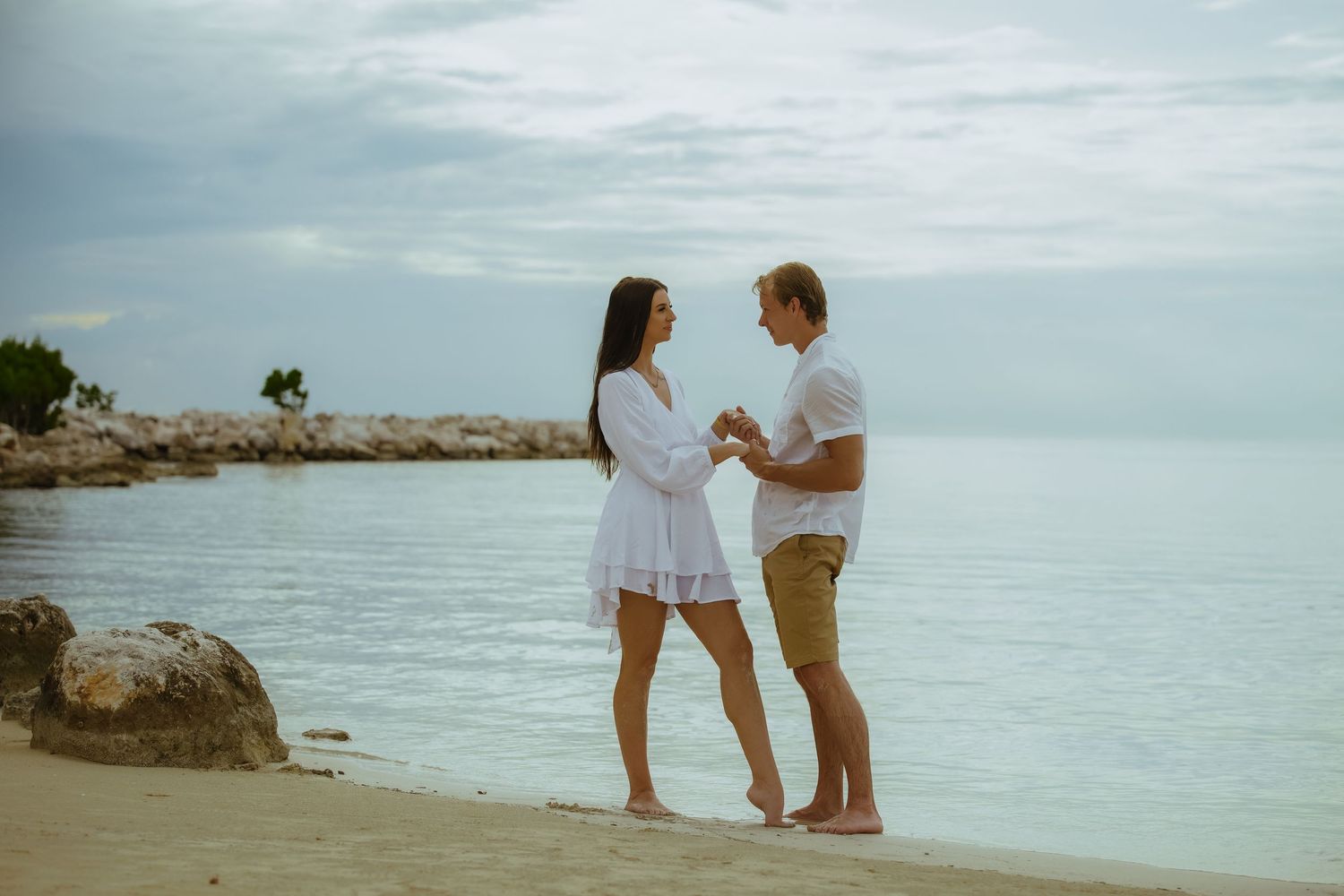 A couple in casual white outfits stands together on a scenic beach with rocky coastline.