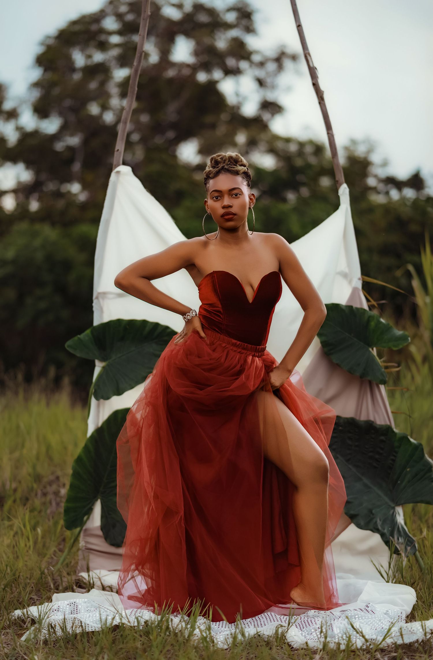 Model in flowing red dress poses dramatically in outdoor garden setting.