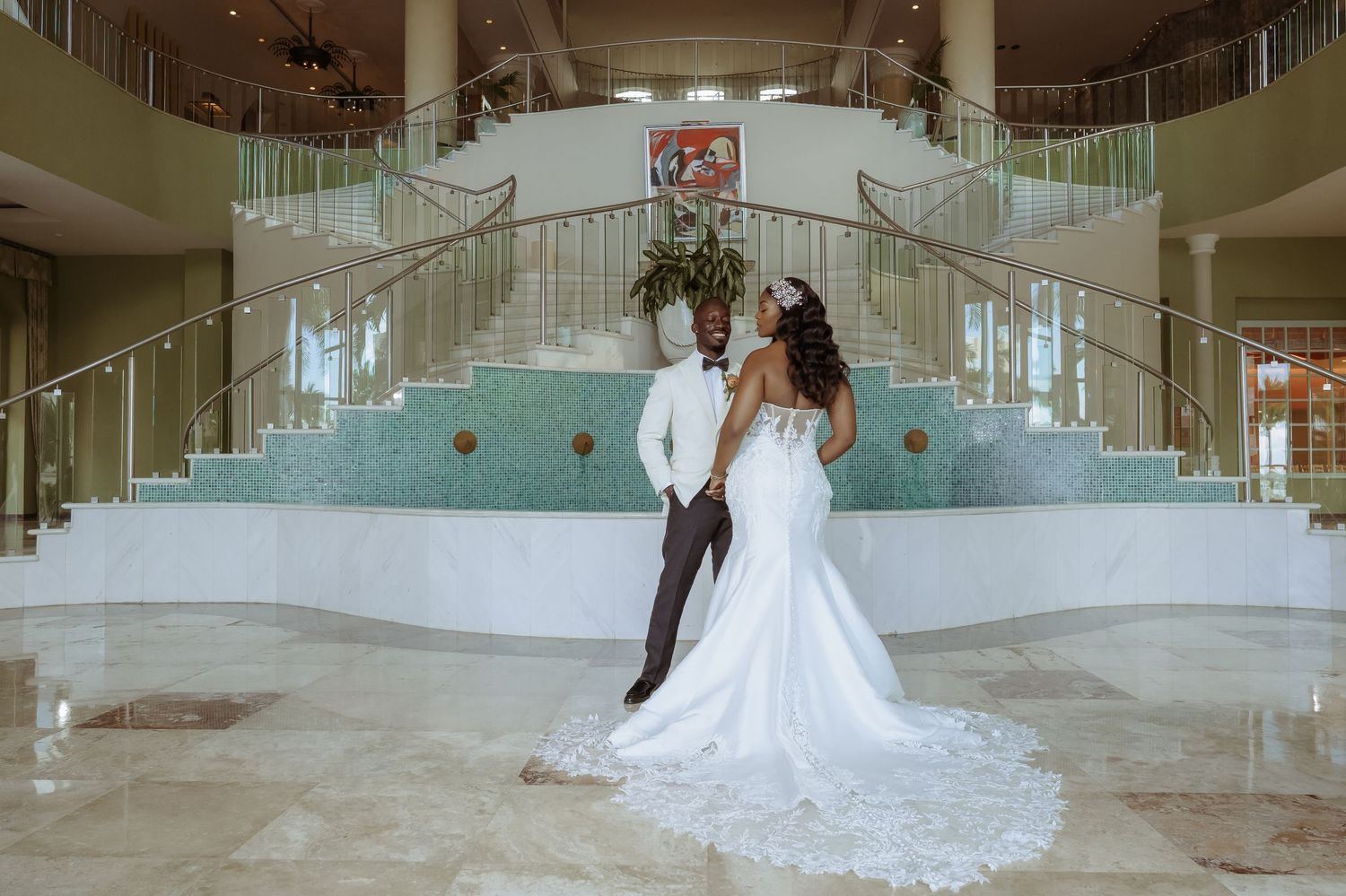 Elegant wedding couple posing in grand foyer with dramatic staircase and fountain.