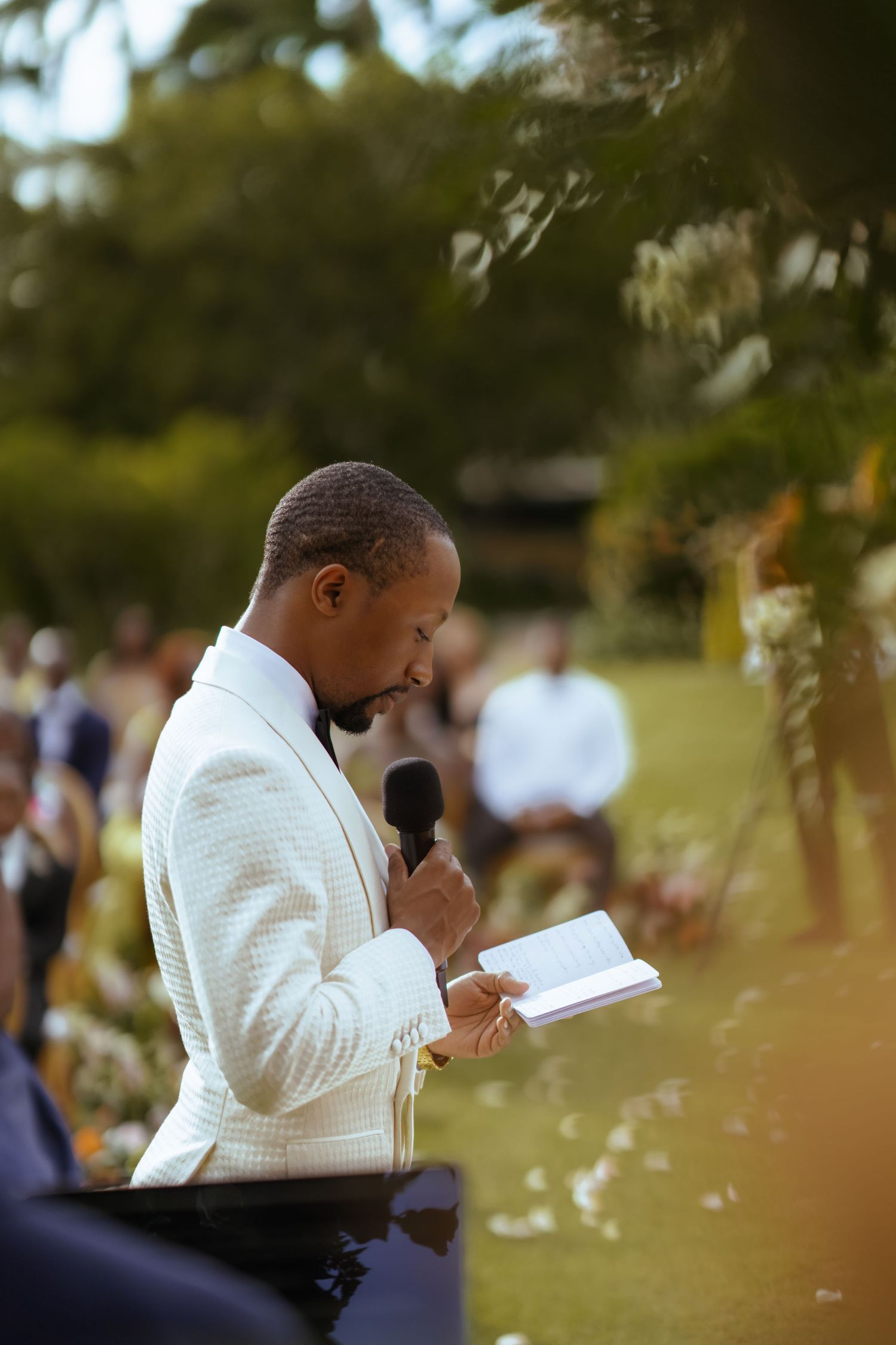 Wedding ceremony in progress with a person in white attire reading from a book outdoors.
