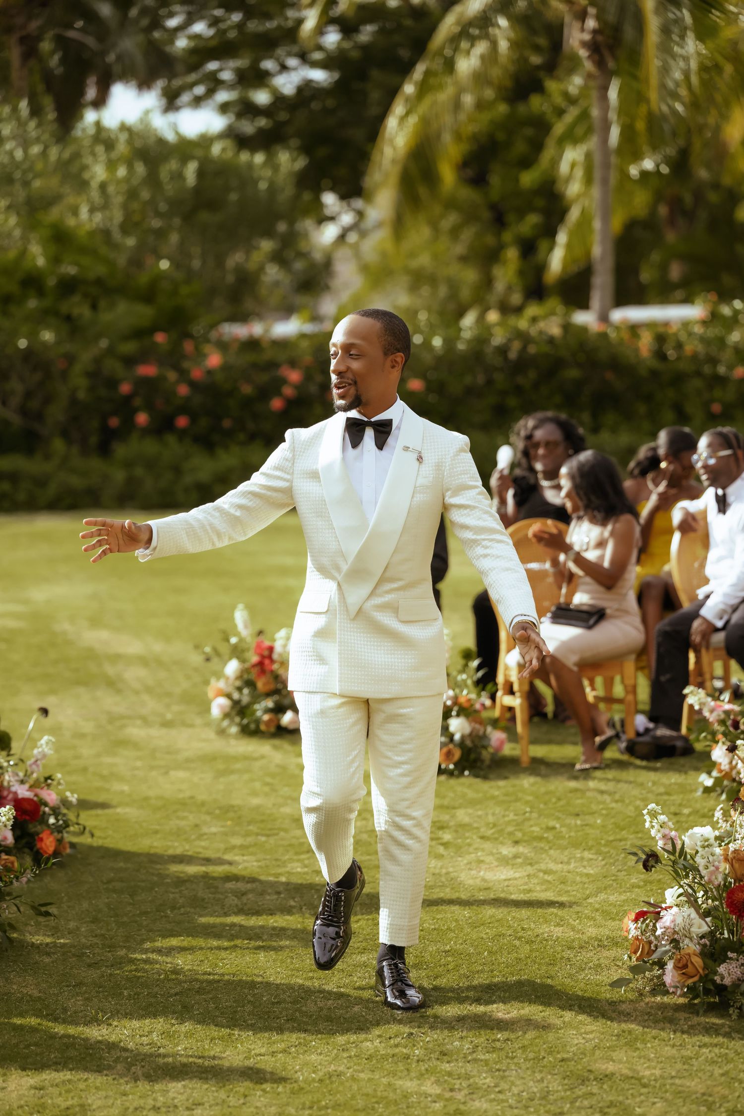 Groom in crisp white tuxedo walking across a lush garden lawn decorated with flowers.