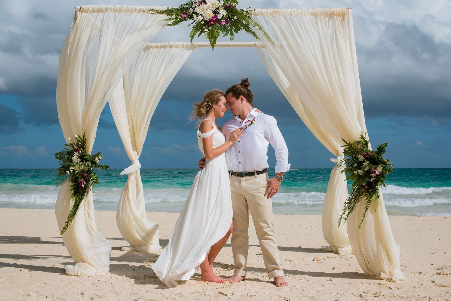 White draped beach wedding arch decorated with flowers stands against turquoise ocean waters.