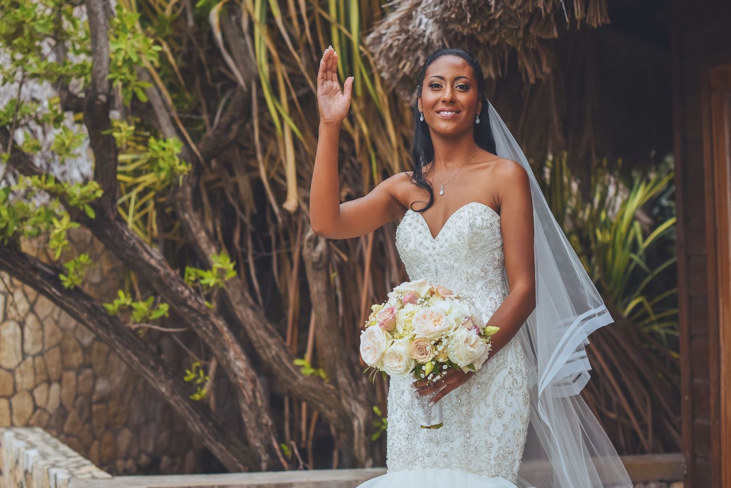 Bride in a detailed white wedding dress poses in a tropical outdoor setting.