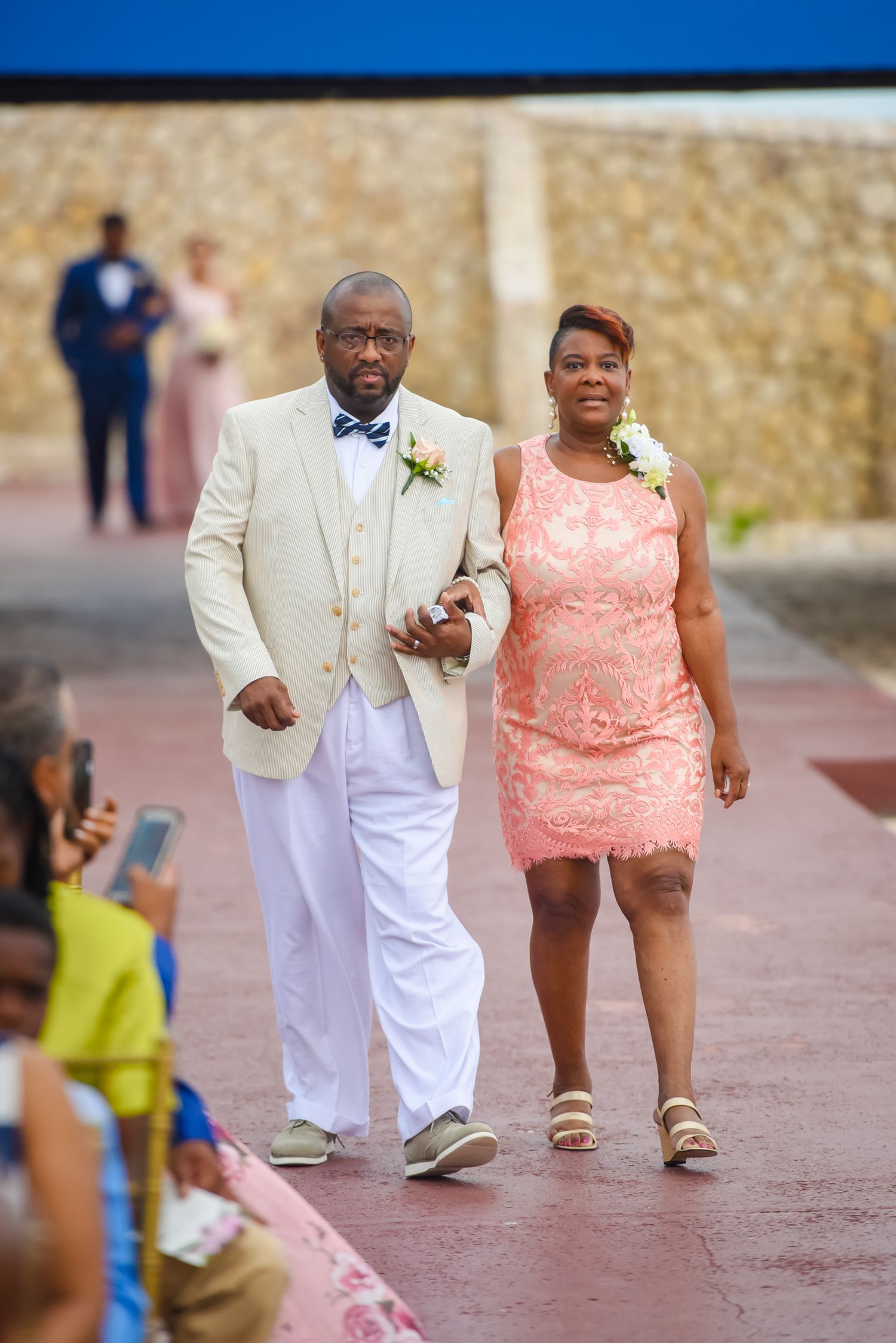 Wedding party members walking aisle in cream suit and coral dress during outdoor ceremony.