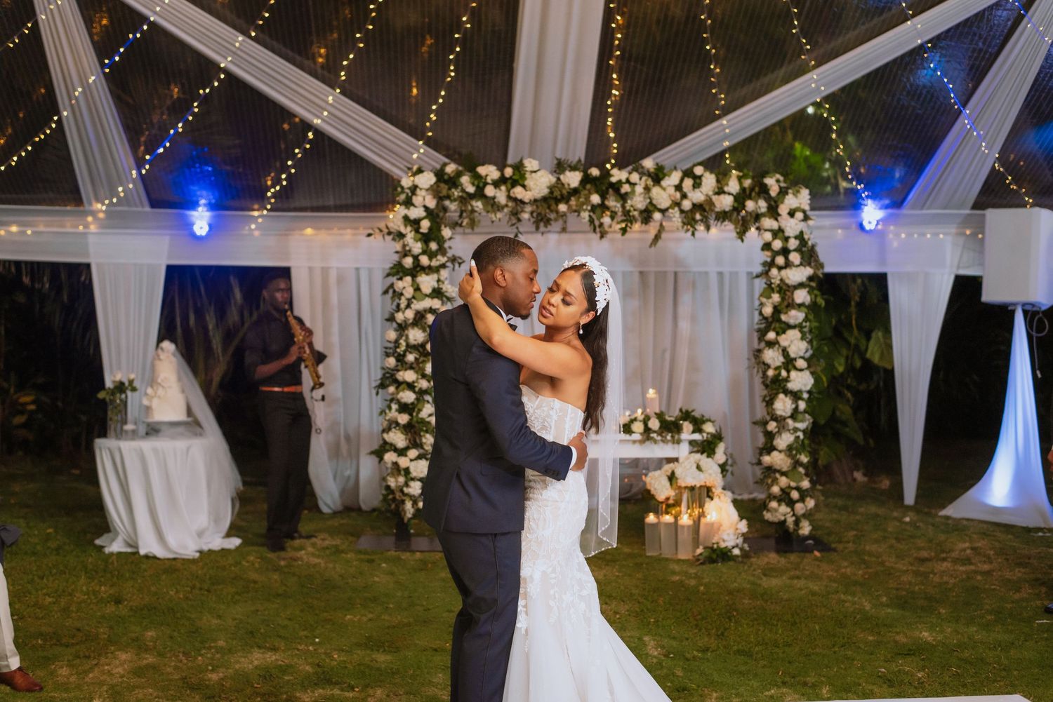 Couple dancing under floral arch with blue uplighting and white drapes at outdoor evening reception.