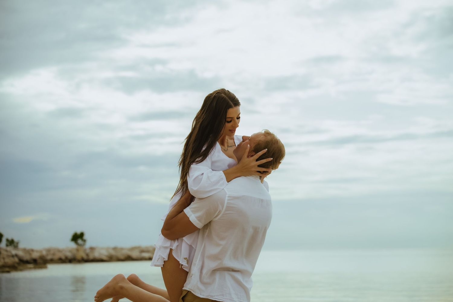 A romantic couple in white clothing shares a joyful moment on a coastal beach.