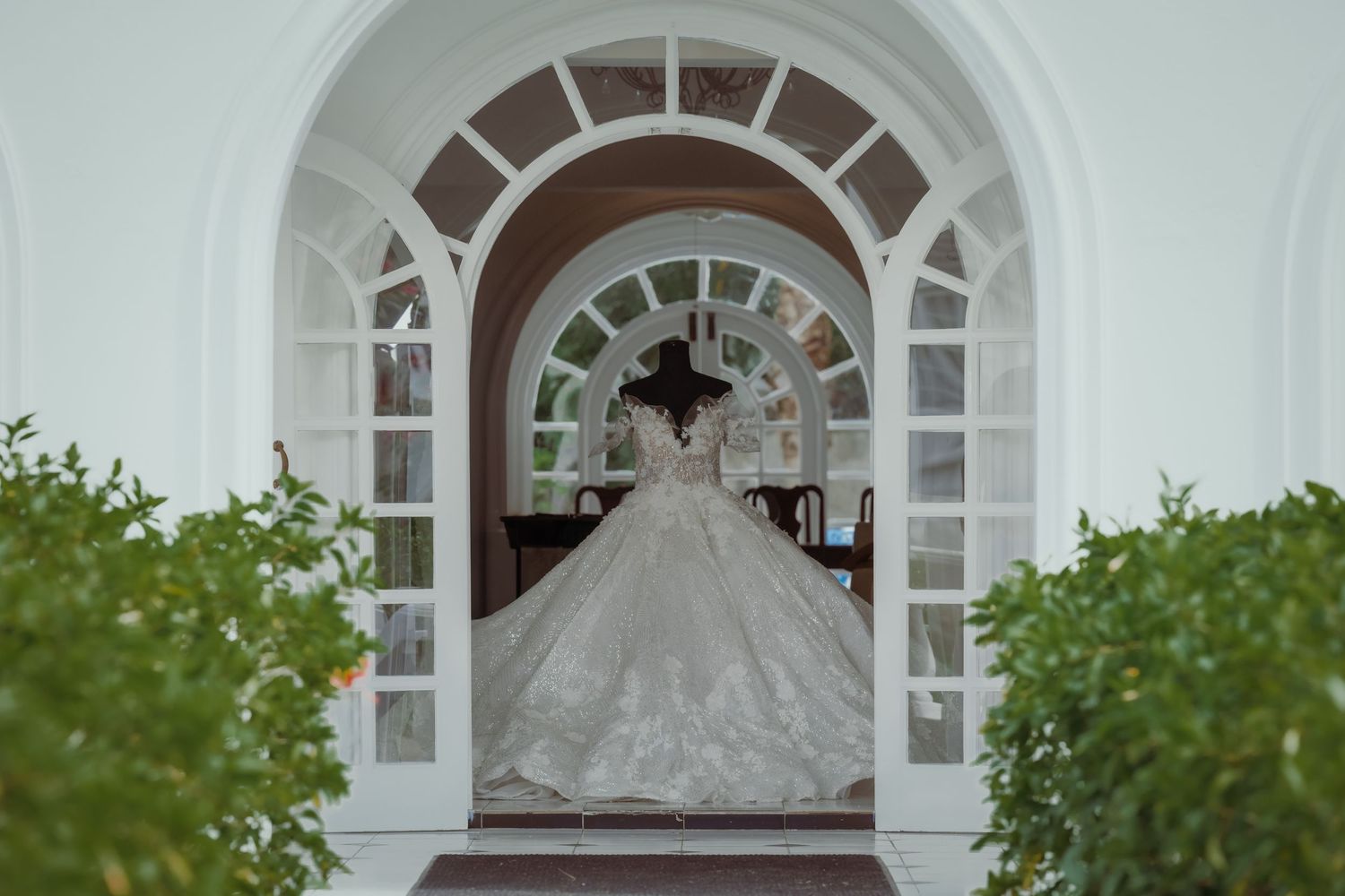 A white wedding dress hangs in an elegant archway with ornate architectural details and lush greenery surrounding it.
