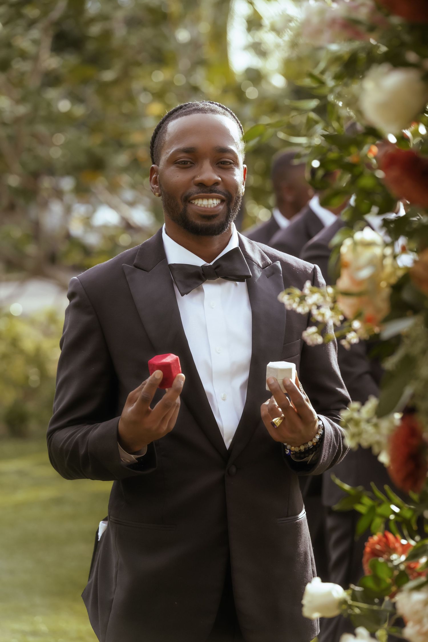 Wedding party member in black tuxedo and bowtie gesturing outdoors.