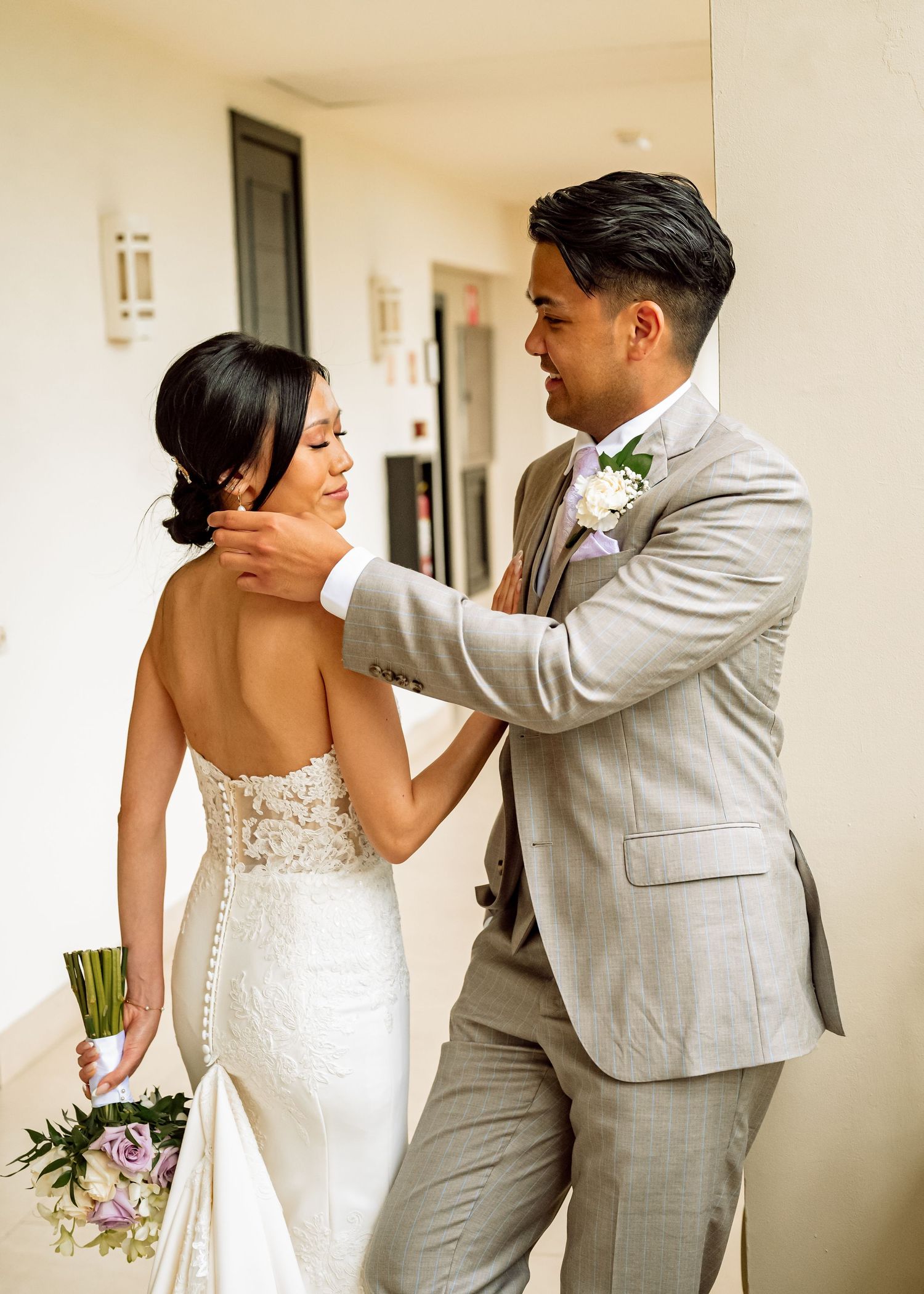 Couple shares intimate first look moment in elegant hallway wearing white and light gray attire.