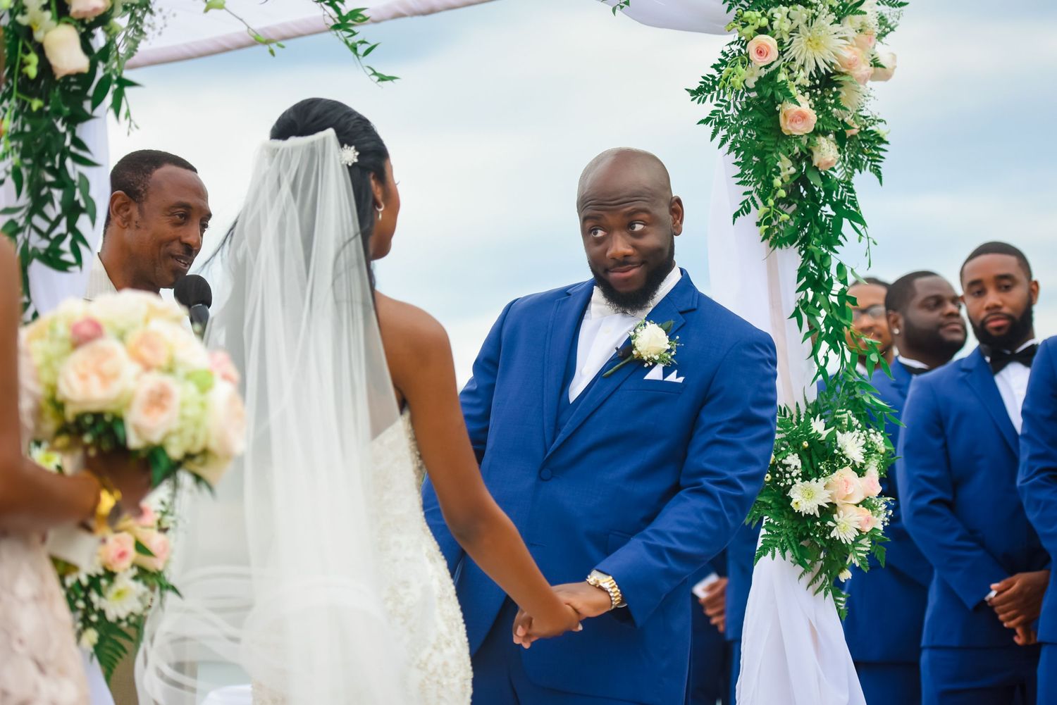 Wedding party stands in formation during ceremony beneath elegant floral arch with white draping.