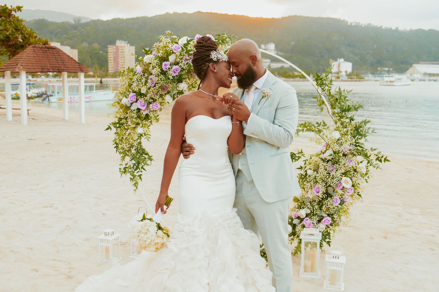 Newlyweds share intimate moment under floral arch on tropical beach at sunset.