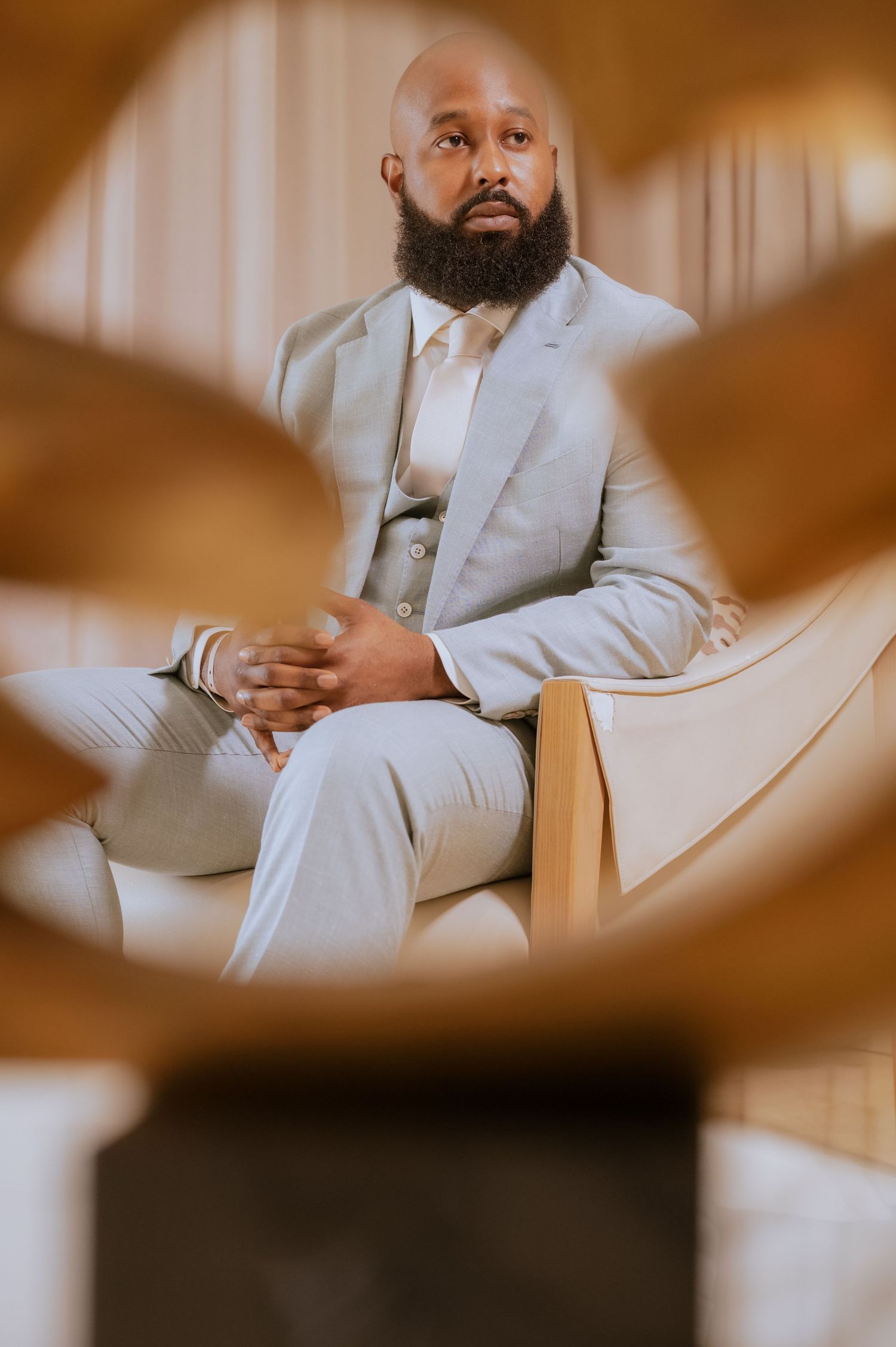 Groom in light gray suit sits thoughtfully on beige couch through artistic framing.