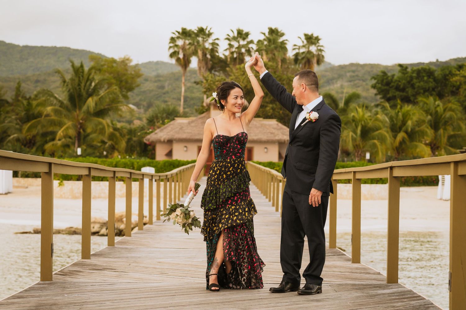 A couple walks along a beachfront boardwalk surrounded by palm trees and tropical scenery.