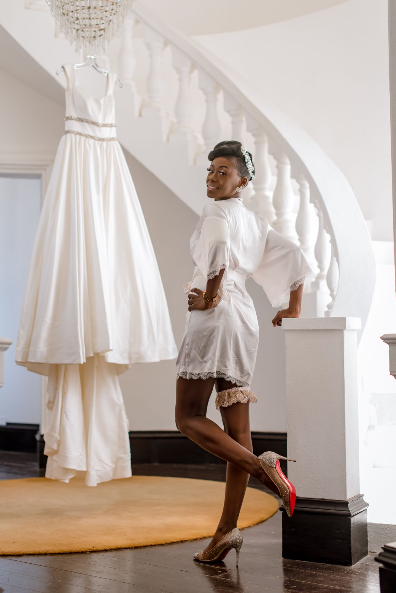 A joyful pose in white bridal preparation attire with red heels near a hanging wedding gown.