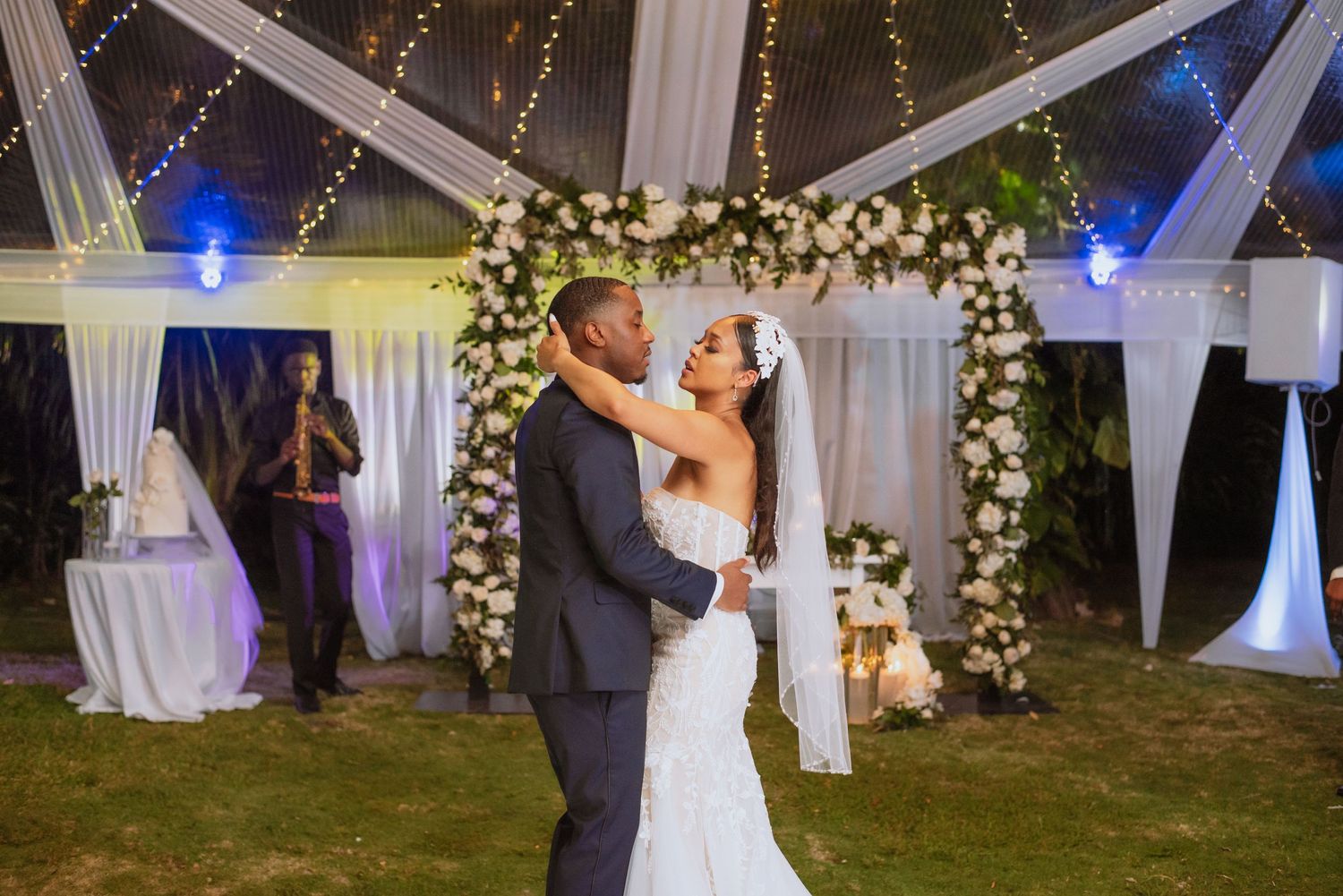 Couple shares romantic first dance under floral arch with twinkling lights in outdoor tent reception setting.