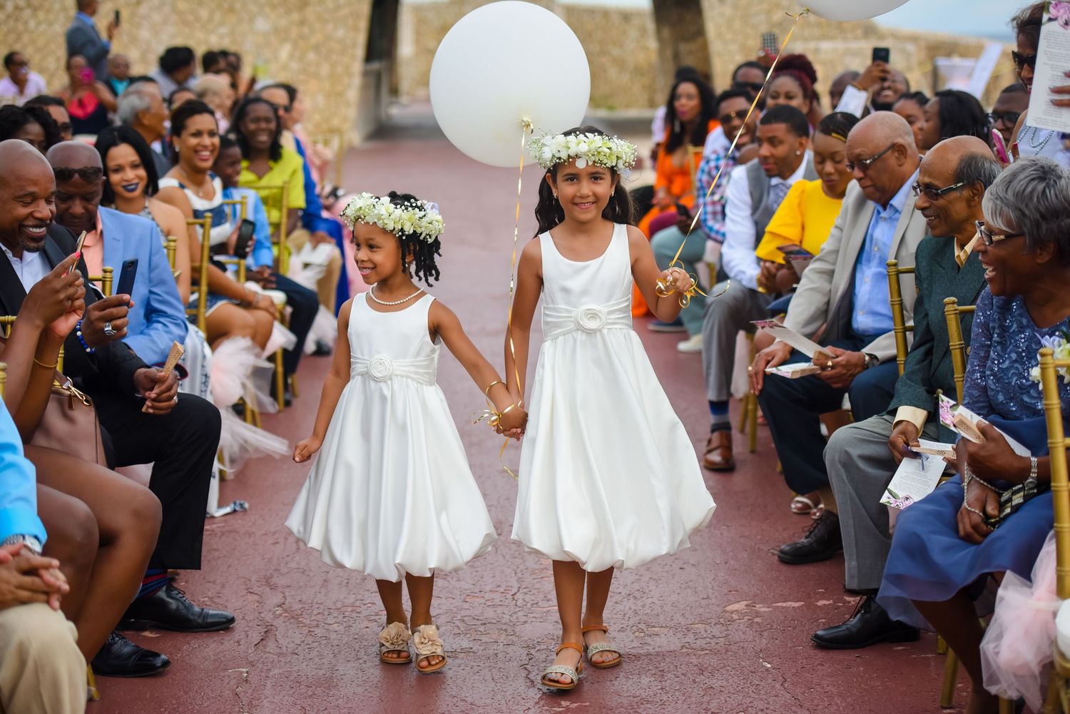 Two young flower girls in white dresses holding hands and balloons during wedding ceremony.