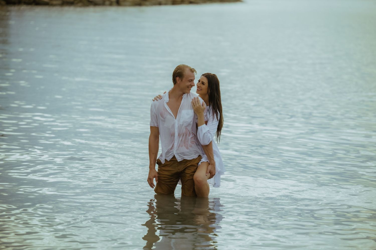 A couple stands together in knee-deep ocean water wearing white shirts.