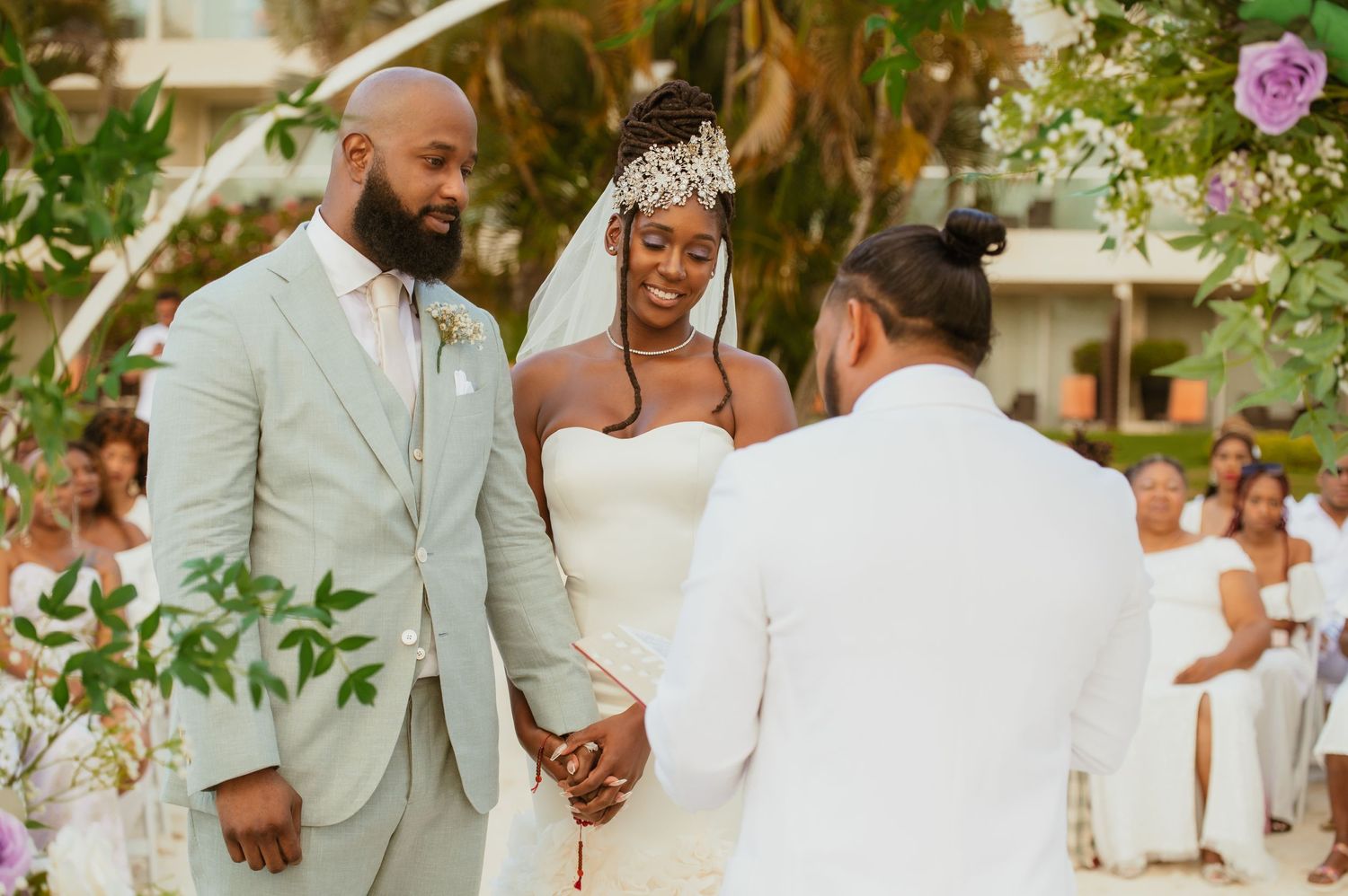 Wedding party in light colored attire gathers for an outdoor celebration surrounded by tropical foliage.