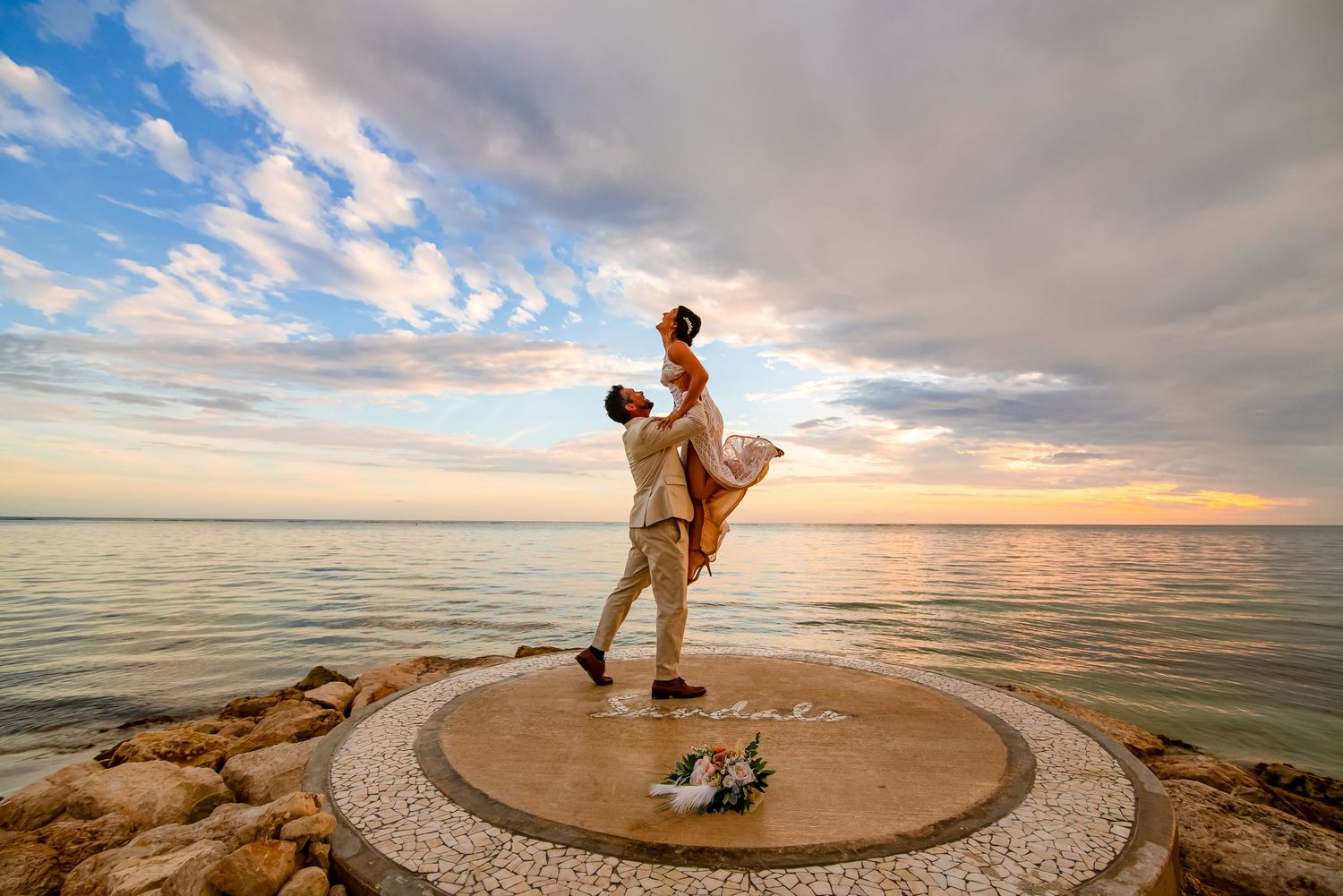 Wedding couple performs romantic dance poses on oceanfront platform at sunset.
