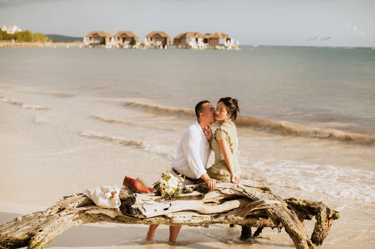 Couple sharing intimate moment on driftwood at beach with overwater bungalows in background.