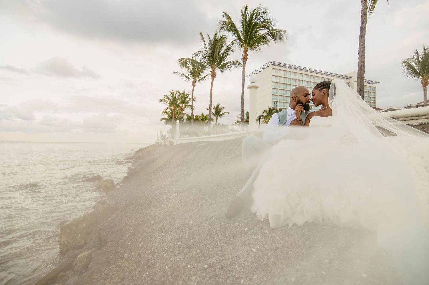 A couple walks on a beach path with palm trees and ocean waves crashing against the shore during sunset.
