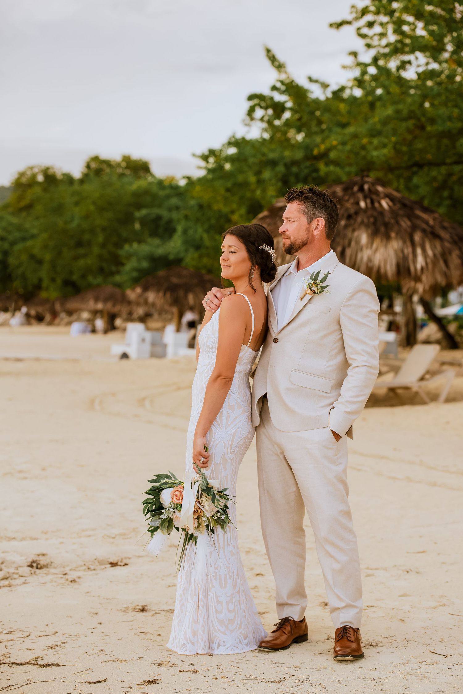 Newlyweds in elegant white outfits pose together on a tropical beach with palm trees.