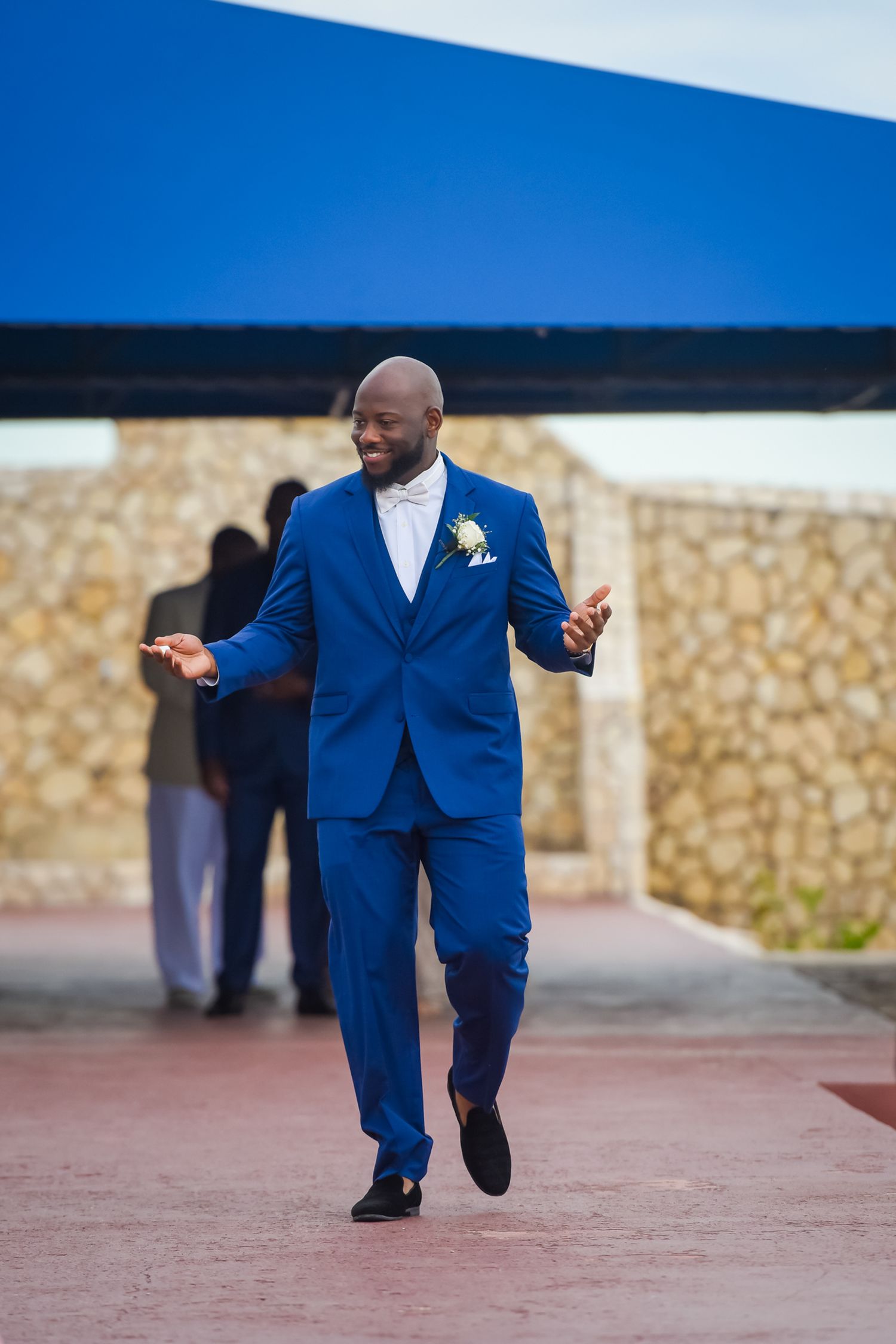 Wedding guest in vibrant blue suit walking and gesturing during outdoor ceremony.
