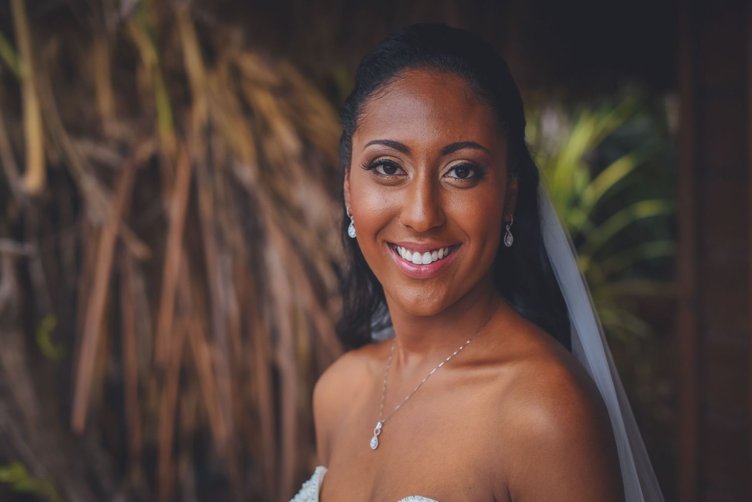 A bride in a white lace wedding dress poses outdoors with a flowing veil against a natural backdrop.