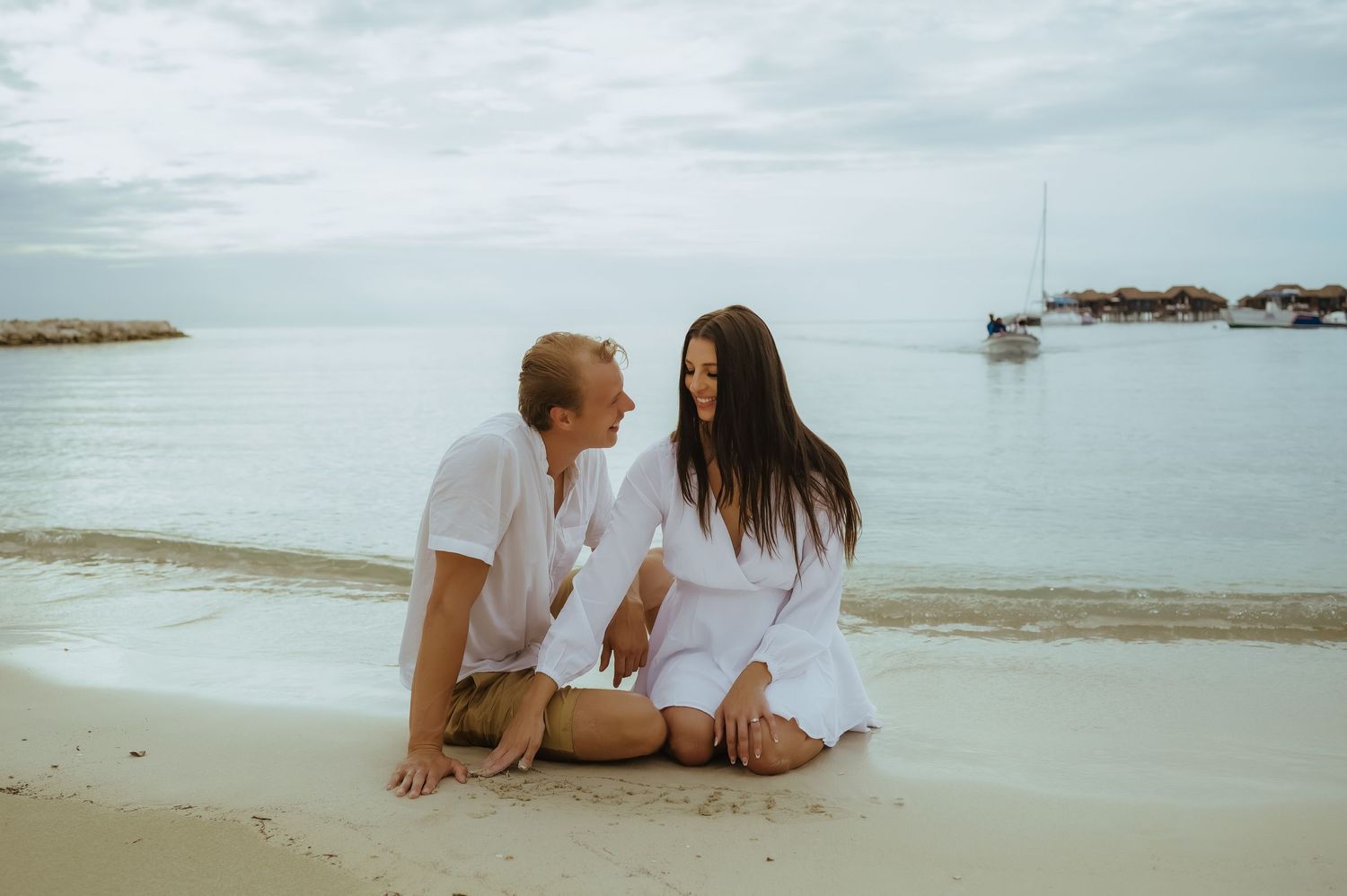 A couple in white clothing sits together on a peaceful beach with boats in the distance.