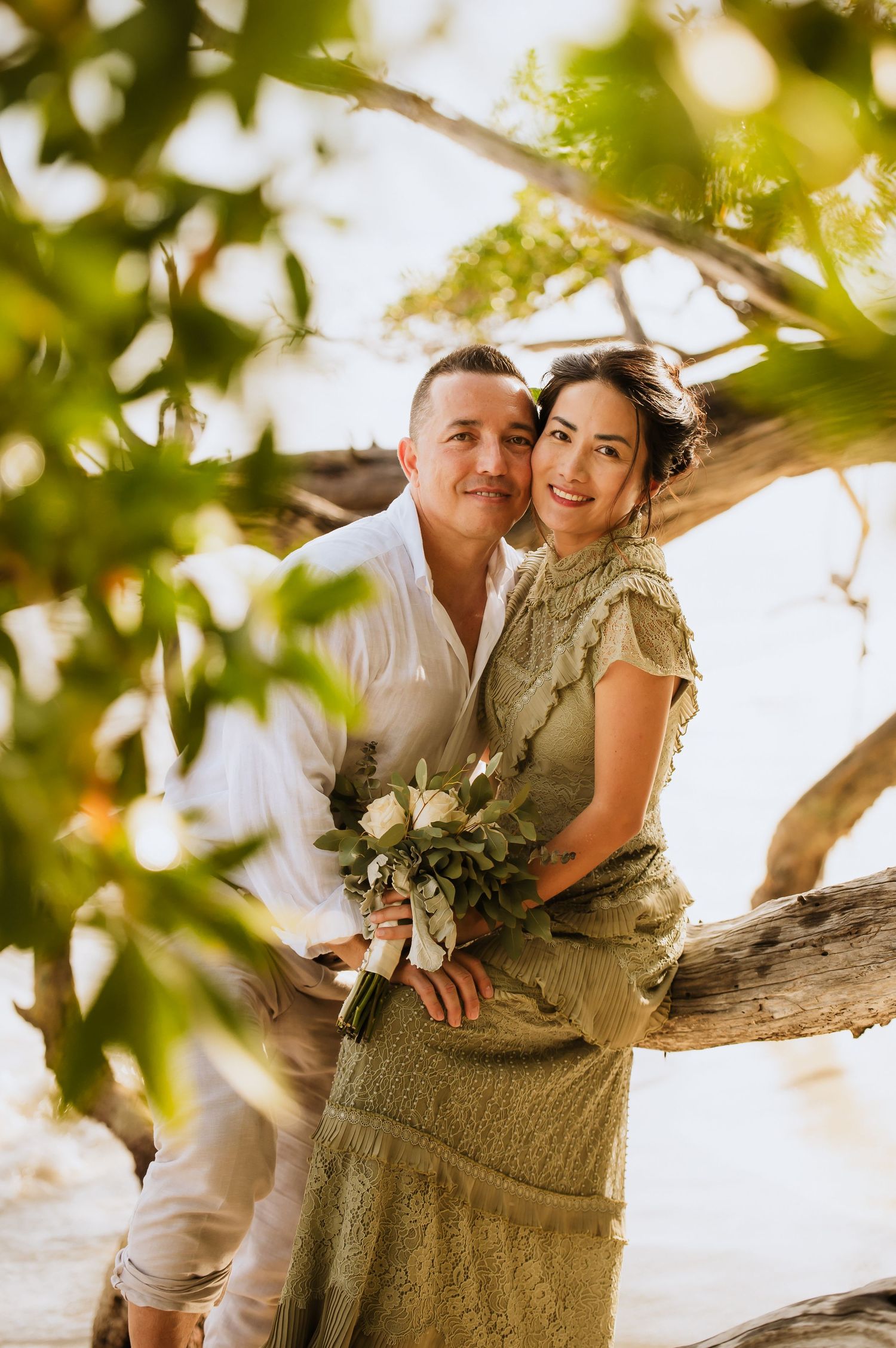 Romantic couple poses among green leaves holding a wedding bouquet.