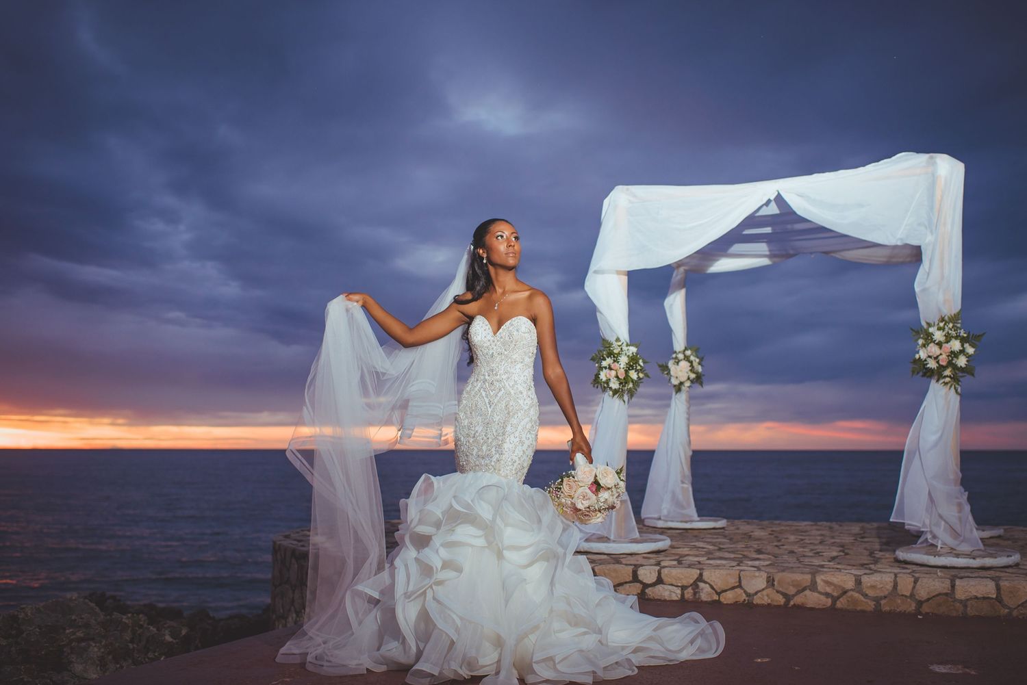 Beach wedding ceremony site with white draped canopy and floral arrangements at sunset.