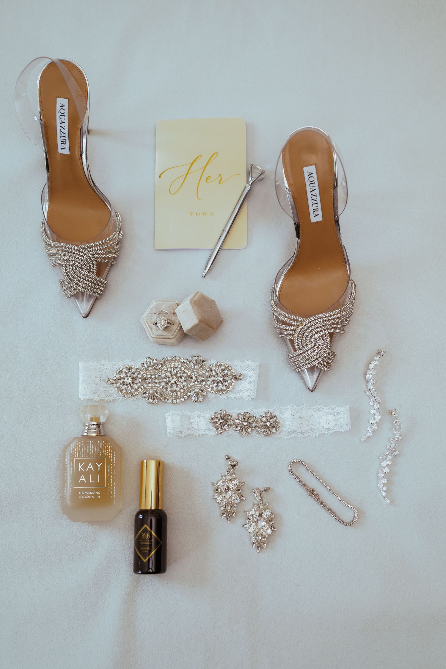 Elegant bridal accessories including silver heels, jewelry, and perfume arranged on white background.