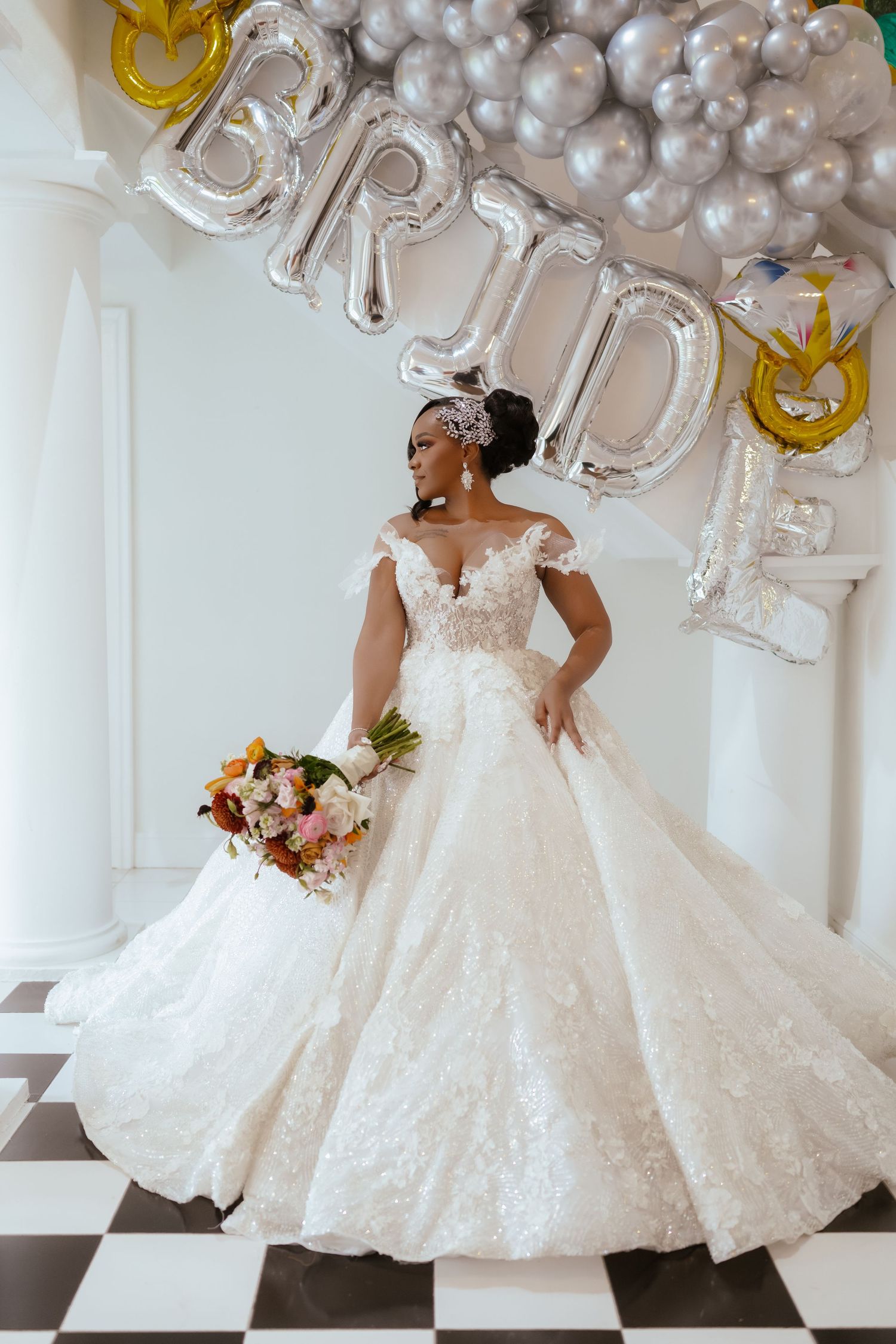 A stunning off-shoulder lace wedding gown with a full skirt photographed against silver balloon decor on a checkered floor.