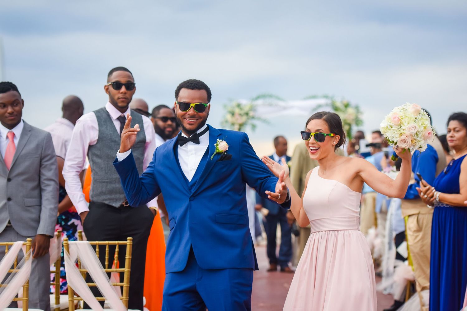 Wedding party members walk down an outdoor aisle with bright smiles and formal attire in blue and pink tones.