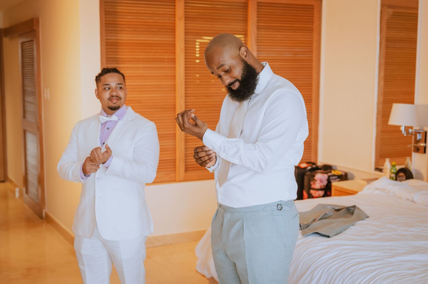 Two men in white and gray outfits adjust their cuffs in hotel room.