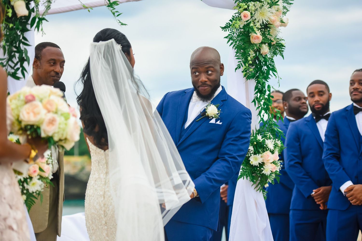 Wedding ceremony by the water with floral arch decorations and guests in formal attire.