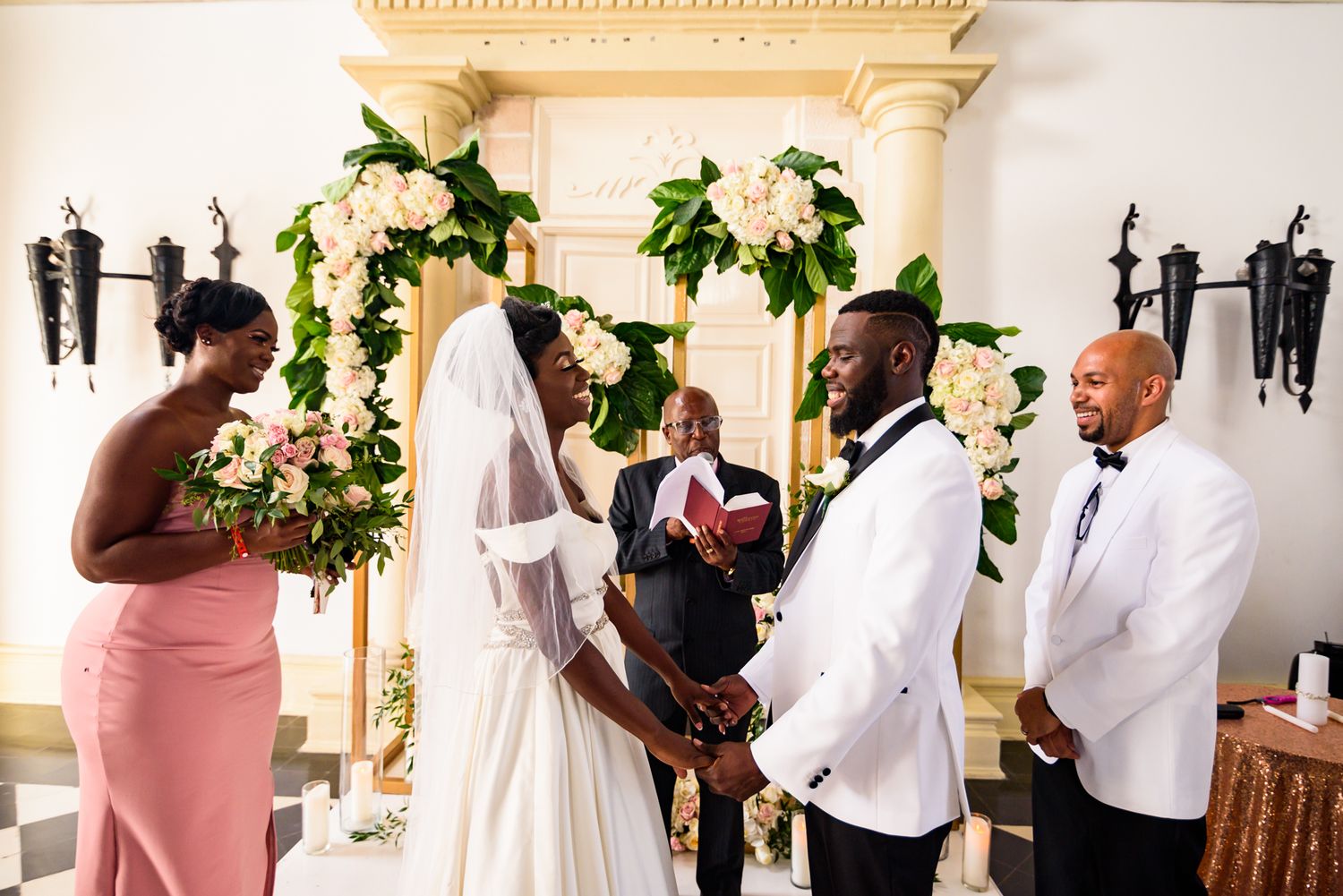 Wedding party in white and pink attire poses together in an ornate indoor venue.