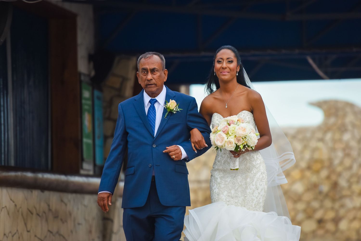Wedding processional on stone-walled walkway with bride carrying white bouquet.
