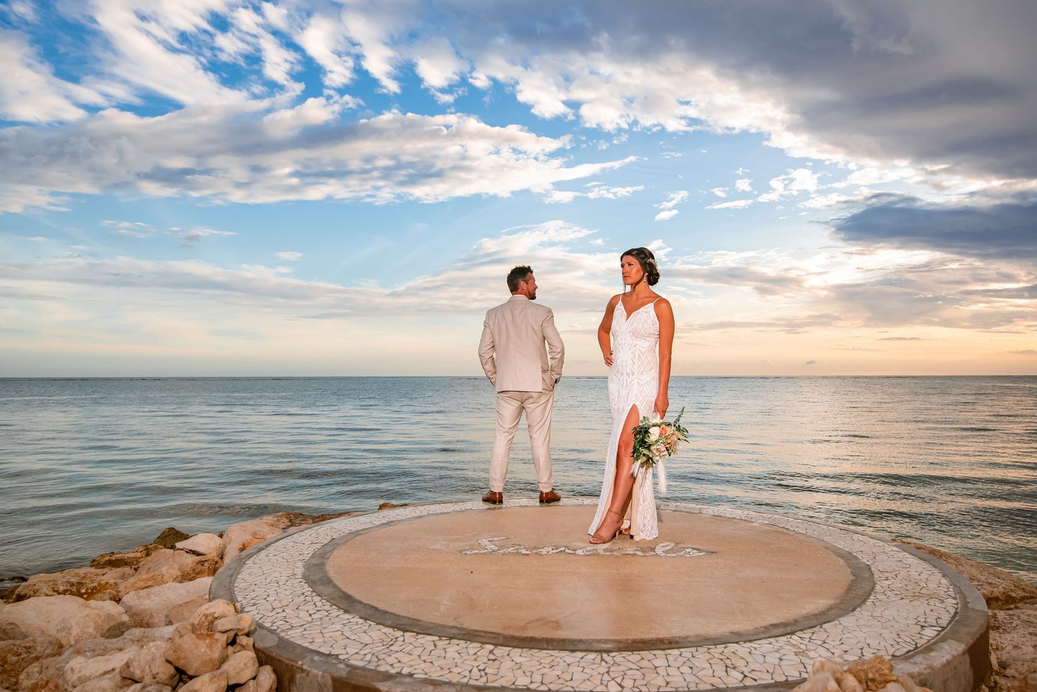 Couple stands on circular platform against dramatic sunset sky and ocean backdrop.