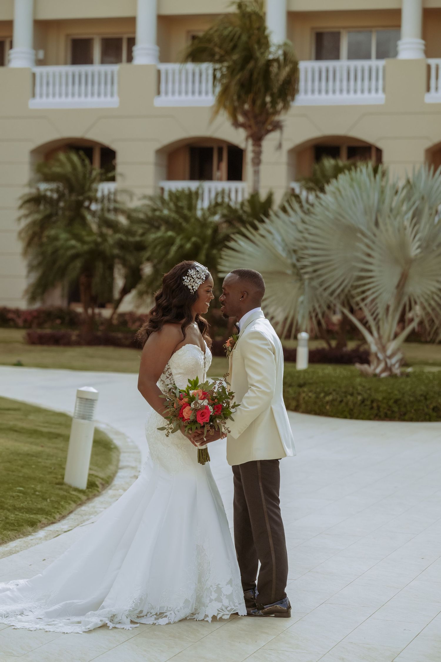 Newlyweds share a romantic moment on a palm-lined path outside a grand building.