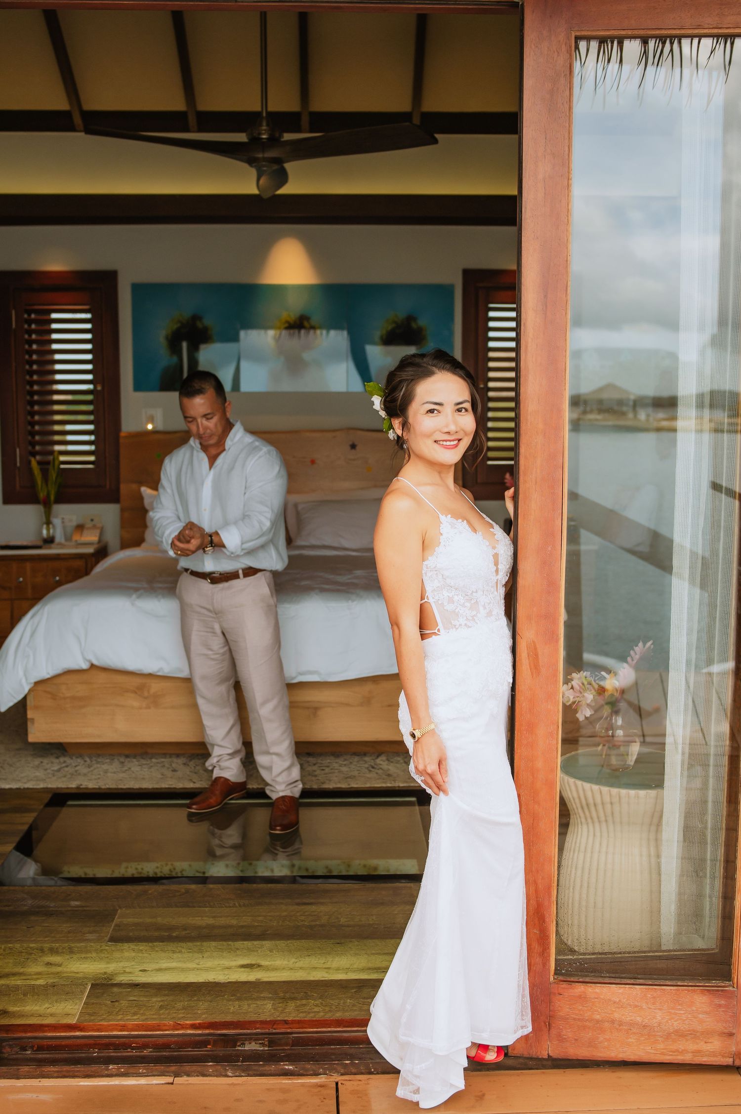 Couple dressed in white poses playfully on a covered outdoor patio with ocean views.