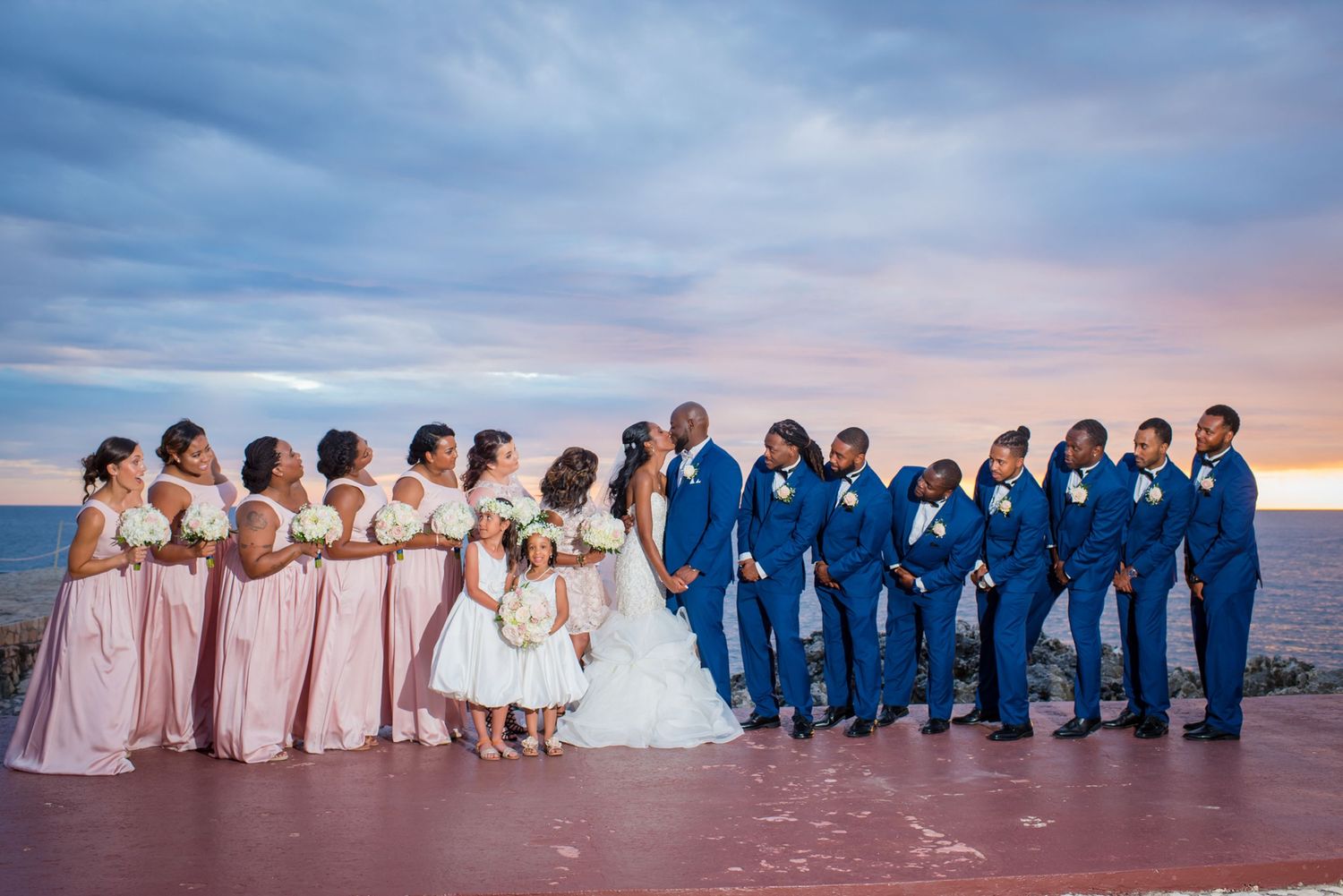 Wedding party members in coordinated blue and pink attire pose against dramatic sunset sky.