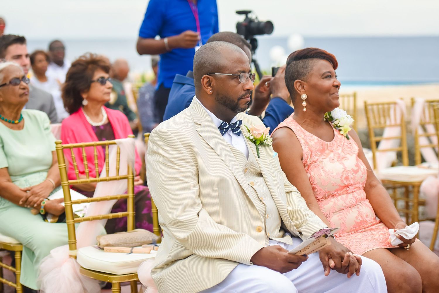 Wedding guests in formal attire seated during an outdoor ceremony.